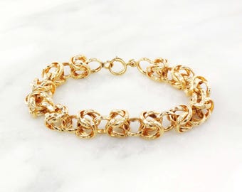 May include: A gold-colored bracelet with a textured, knotted design. The bracelet features a series of interconnected, rounded links, creating a chain-like appearance. The clasp is a simple, circular design. The bracelet is displayed on a white surface.