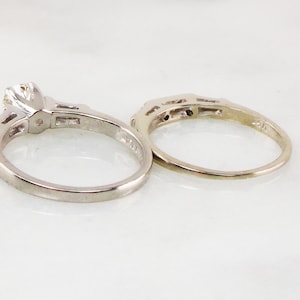 May include: A set of two silver engagement rings. The larger ring has a single round diamond in the center and smaller diamonds on the band. The smaller ring has a band of smaller diamonds.