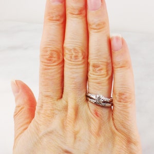 May include: A close-up of a hand wearing a silver wedding band and a diamond engagement ring.