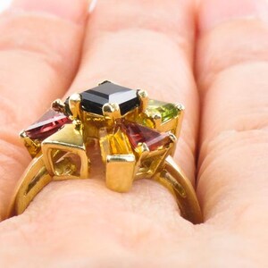 Op de afbeelding: Een gouden ring met een zwarte vierkante edelsteen omringd door kleinere rode, groene en gele edelstenen.
