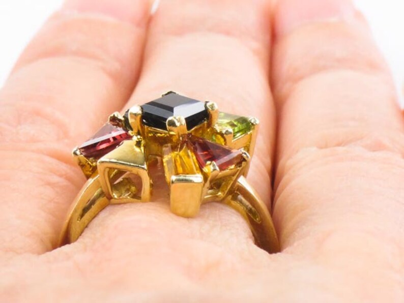 Op de afbeelding: Een gouden ring met een zwarte vierkante edelsteen omringd door kleinere rode, groene en gele edelstenen.