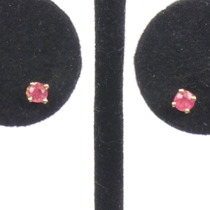 May include: A pair of stud earrings featuring small, round, faceted pink gemstones set in gold-toned metal prongs. Each earring is centered on a circular black velvet display pad, showcasing the vibrant pink color of the stones against the dark background.