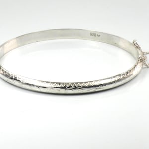 May include: A silver bangle bracelet with a decorative etched pattern. The bracelet has a hinged clasp and a safety chain. The metal is bright and reflective, and the design is simple and elegant. The bracelet is a piece of jewelry.