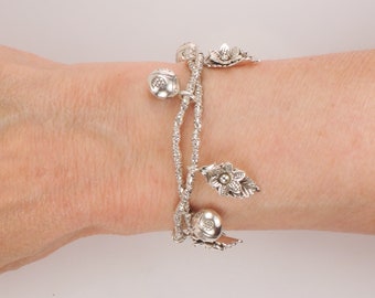 May include: Silver charm bracelet with multiple charms, including flowers and leaves. The bracelet is on a person's wrist.