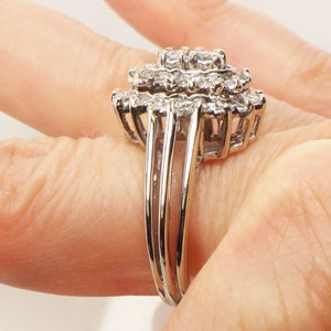 Op de afbeelding: Een zilveren ring met een grote cluster van kleine diamanten, gezet in een bloemvorm. De ring heeft een drievoudig bandontwerp.