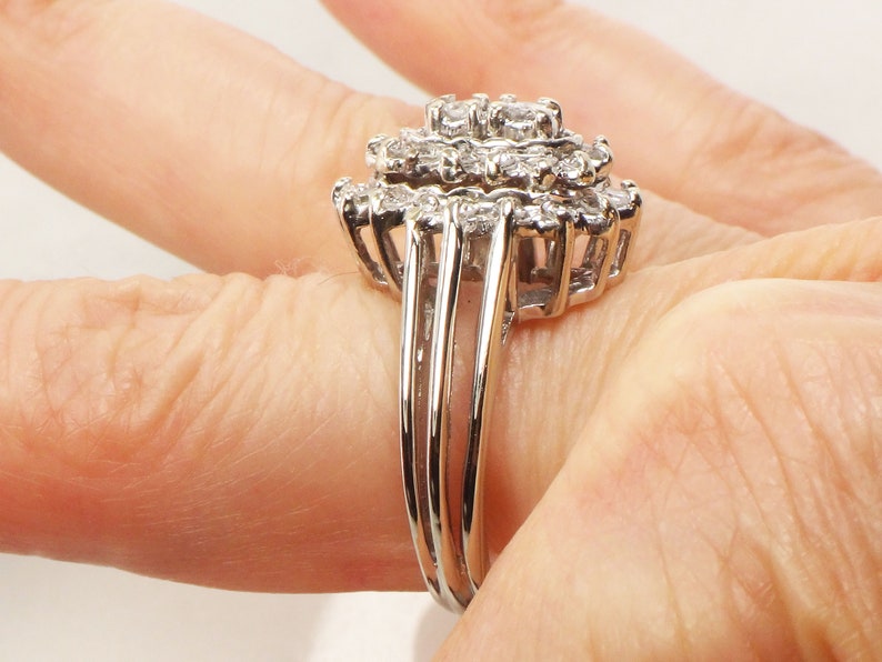 Op de afbeelding: Een zilveren ring met een grote cluster van kleine diamanten, gezet in een bloemvorm. De ring heeft een drievoudig bandontwerp.