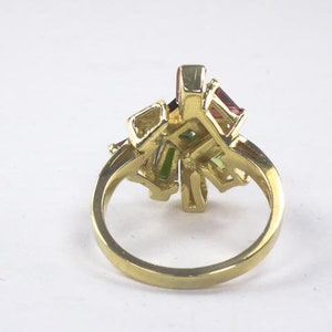 Op de afbeelding: Een gouden ring met een cluster van kleurrijke edelstenen. De ring heeft een delicaat, ingewikkeld ontwerp en bevat een verscheidenheid aan kleuren, waaronder roze, groen en rood.
