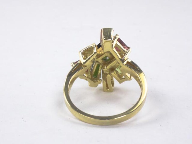 Op de afbeelding: Een gouden ring met een cluster van kleurrijke edelstenen. De ring heeft een delicaat, ingewikkeld ontwerp en bevat een verscheidenheid aan kleuren, waaronder roze, groen en rood.