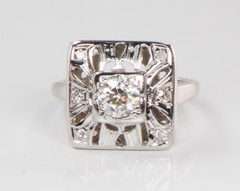 May include: A silver ring with a square-shaped setting featuring a large, clear center stone surrounded by smaller diamonds. The ring has a delicate, intricate design.