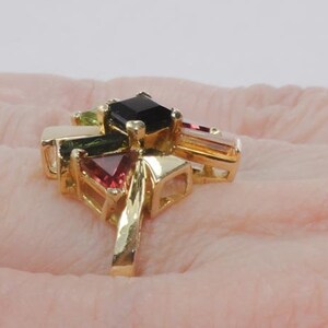 Op de afbeelding: Een gouden ring met een cluster van gekleurde edelstenen. De ring heeft een zwarte vierkante steen in het midden, omringd door groene, roze en heldere stenen.