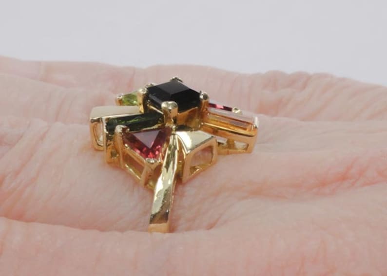 Op de afbeelding: Een gouden ring met een cluster van gekleurde edelstenen. De ring heeft een zwarte vierkante steen in het midden, omringd door groene, roze en heldere stenen.
