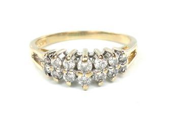 May include: A gold-tone ring featuring a cluster of small, round, clear gemstones. The ring has a split shank design and a series of gemstones set in a floral-like pattern. The ring is set against a white background.
