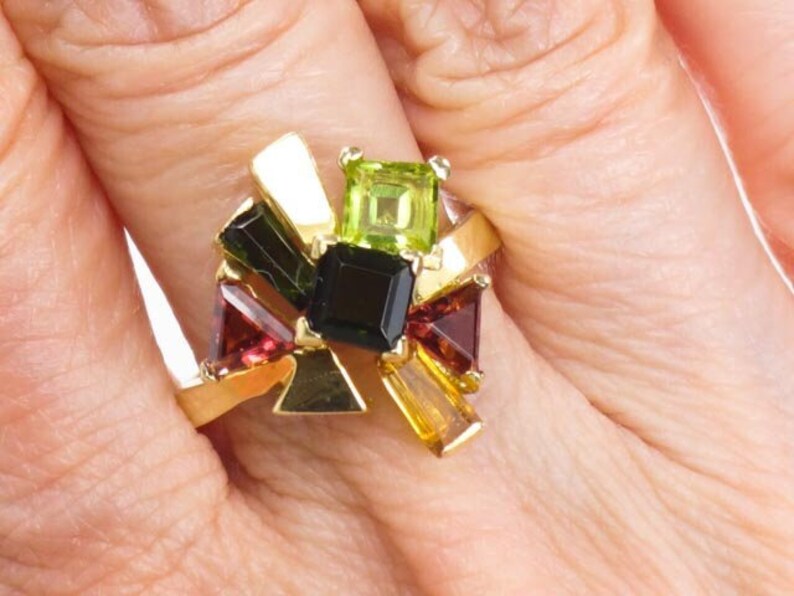 Op de afbeelding: Een gouden ring met een cluster van kleurrijke edelstenen, waaronder groen, zwart en rood.