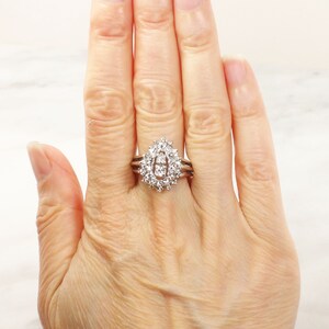 Op de afbeelding: Een zilveren ring met een grote ovale middelste steen omringd door kleinere diamanten. De ring wordt gedragen aan een vrouwenvinger.