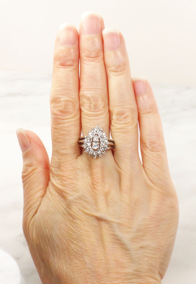 Op de afbeelding: Een zilveren ring met een grote ovale middelste steen omringd door kleinere diamanten. De ring wordt gedragen aan een vrouwenvinger.