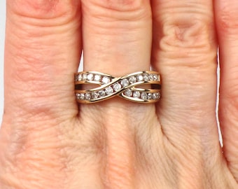 May include: A gold-toned ring with an infinity design, set with multiple small, clear gemstones. The ring is worn on a finger, showcasing its intricate, interwoven bands and sparkling stones.
