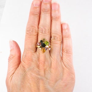 Op de afbeelding: Een gouden ring met een cluster van veelkleurige edelstenen. De edelstenen zijn in een stervormig patroon gerangschikt en omvatten groen, zwart, paars en geel.