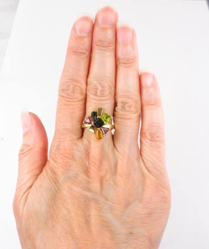 Op de afbeelding: Een gouden ring met een cluster van veelkleurige edelstenen. De edelstenen zijn in een stervormig patroon gerangschikt en omvatten groen, zwart, paars en geel.