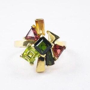 Op de afbeelding: Een gouden ring met een cluster van groene, rode en oranje edelstenen. De ring heeft een verscheidenheid aan vormen en maten edelstenen, wat een uniek en opvallend ontwerp cre&euml;ert.