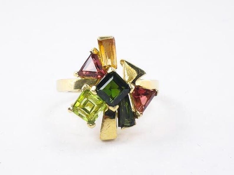 Op de afbeelding: Een gouden ring met een cluster van groene, rode en oranje edelstenen. De ring heeft een verscheidenheid aan vormen en maten edelstenen, wat een uniek en opvallend ontwerp cre&euml;ert.