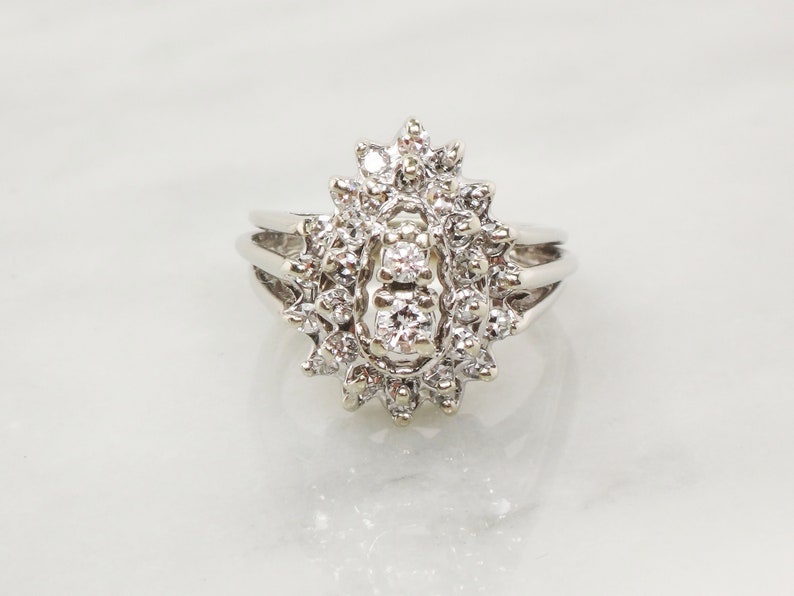 Op de afbeelding: Een zilveren ring met een grote, ovale cluster van diamanten. De ring heeft een delicate band en een middelste steen die omringd is door kleinere diamanten.
