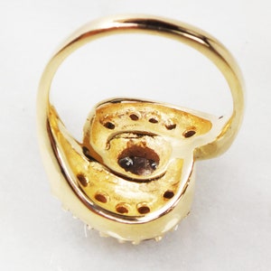 May include: A gold ring with a large, round, clear stone set in the center. The stone is surrounded by smaller, clear stones. The ring is set on a white background.