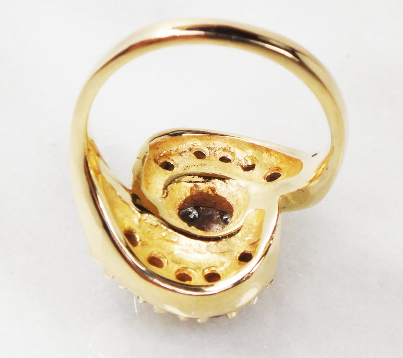 May include: A gold ring with a large, round, clear stone set in the center. The stone is surrounded by smaller, clear stones. The ring is set on a white background.