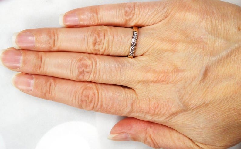 May include: A close-up of a hand wearing a thin gold band with small clear stones set in the metal.