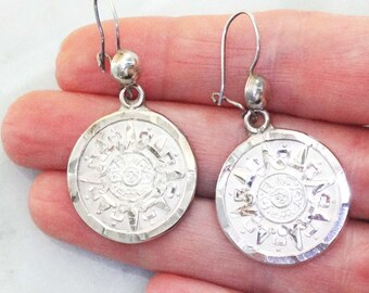 May include: Silver dangle earrings with a circular design featuring a stylized sun with intricate details. The earrings have a hammered texture and a hook closure.