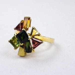 Op de afbeelding: Een gouden ring met een cluster van kleurrijke edelstenen. De ring heeft een verscheidenheid aan groene, rode en gele stenen in verschillende vormen en maten.