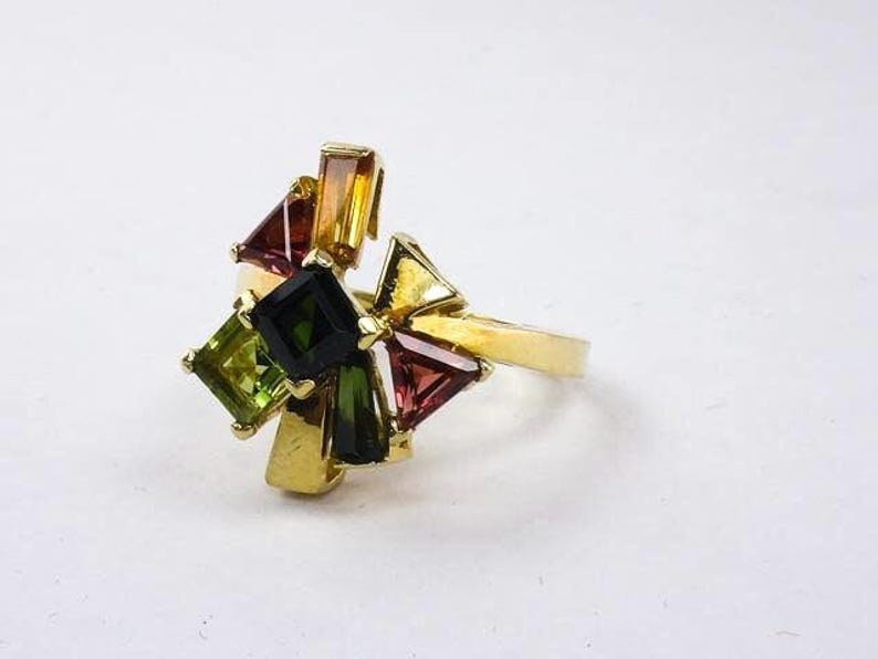 Op de afbeelding: Een gouden ring met een cluster van kleurrijke edelstenen. De ring heeft een verscheidenheid aan groene, rode en gele stenen in verschillende vormen en maten.