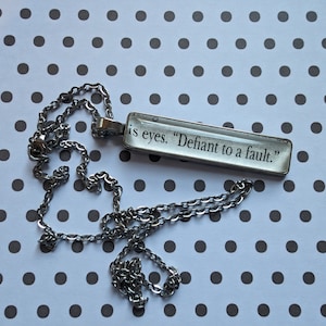 Puede incluir: Un collar de cadena plateada con un colgante rectangular de vidrio que dice "is eyes. Defiant to a fault."
