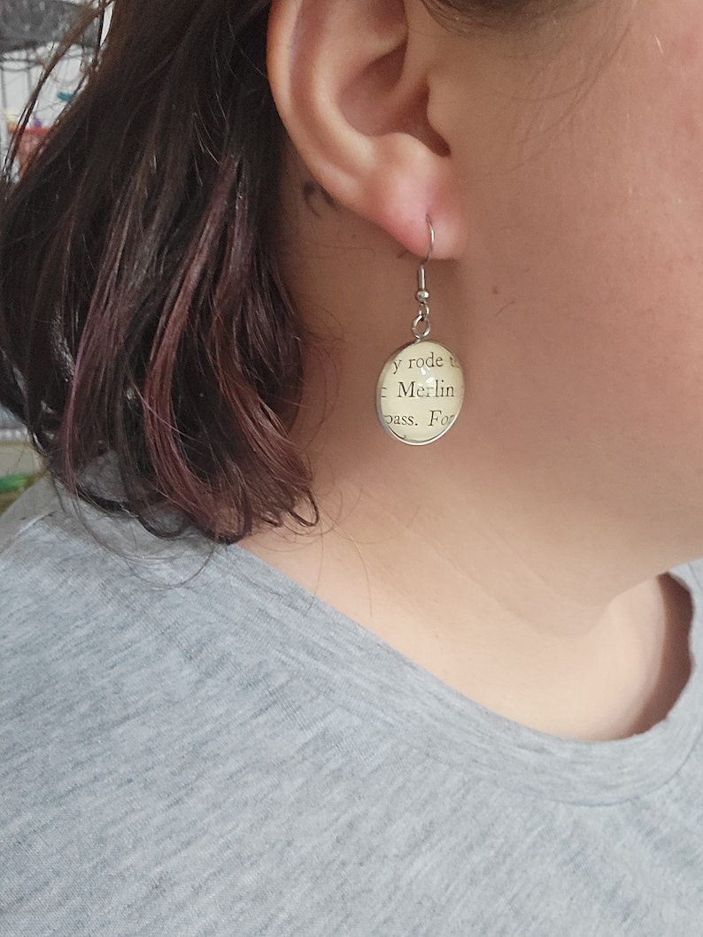 Op de afbeelding: Zilveren oorbellen met een ronde, cr&egrave;mekleurige hanger met de tekst "y rode to - Merlin pass. For"