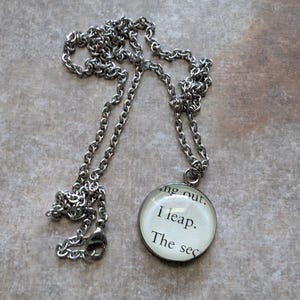 Puede incluir: Collar plateado con cadena y colgante redondo. El colgante muestra texto de una página de libro: "I leap. The sec..." El collar se muestra sobre una superficie texturizada.