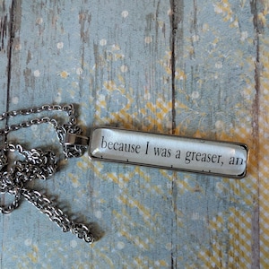 May include: A silver-colored chain necklace with a rectangular pendant. The pendant has the text "because I was a greaser, an" in black font on a white background. The necklace rests on a blue and yellow patterned surface.