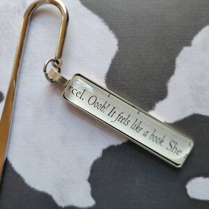 May include: Silver bookmark with a glass charm that reads "Oooo! It feels like a book She", a popular quote from the book "The Book Thief" by Markus Zusak.