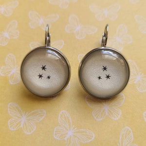 May include: A pair of silver earrings with a white background and a black constellation design. The earrings are round and have a silver hook.