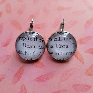 May include: A pair of silver dangle earrings with glass round charms. The charms have a white background with black text that reads "espite the Dean tak mischief. A bell" and "call me Cora. D y in torme tod".