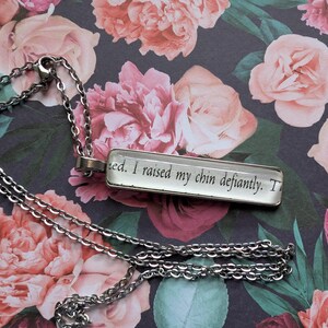 May include: A silver-toned necklace with a rectangular pendant. The pendant has a clear surface with the text "I raised my chin defiantly." The background features a floral pattern with pink and red roses on a dark background.