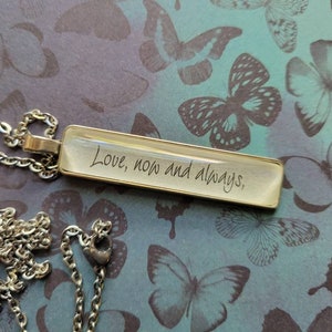 May include: A silver chain necklace with a rectangular pendant that says "Love, now and always". The pendant is clear and has a silver border. The necklace is on a background of blue and purple butterflies.