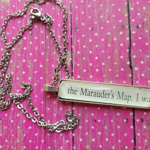 May include: A silver chain necklace with a rectangular pendant featuring the text "the Marauder's Map. I was", printed in black on a white background.