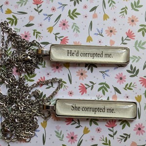 May include: Two silver necklaces with glass pendants. The pendants are rectangular and have white backgrounds with black text. The top pendant says "He'd corrupted me." The bottom pendant says "She corrupted me."