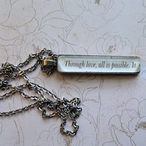 Puede incluir: Collar plateado con un colgante rectangular. El colgante presenta la cita "Through love, all is possible. It" en texto negro sobre fondo blanco. La cadena es de estilo clásico.