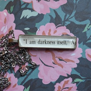 Puede incluir: Un collar de plata con un colgante rectangular que presenta el texto "Soy la oscuridad misma, A". El colgante está sobre un fondo floral.