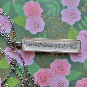 Puede incluir: Un collar de cadena de plata con un colgante rectangular de vidrio que dice "The Seven Husbands of Evelyn Hugo".
