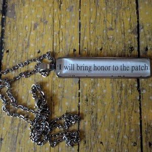 Puede incluir: Un collar de cadena plateada con un colgante rectangular. El colgante tiene el texto "I will bring honor to the patch" en negro sobre un fondo blanco. El collar descansa sobre una superficie de madera amarilla envejecida.