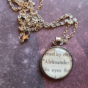 Puede incluir: Un collar de cadena de plata con un colgante redondo de vidrio que presenta un extracto impreso de un libro. El texto dice "ened by eter "Aleksander." His eyes flu"