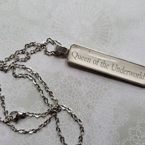 May include: A silver chain necklace with a rectangular glass pendant that says "Queen of the Underworld!"