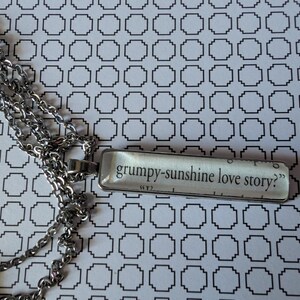 May include: Silver-toned necklace with a rectangular pendant. The pendant features the text "grumpy-sunshine love story?" in a black, serif font. The chain is made of small, interconnected links. The background is a white surface with a black geometric pattern.