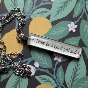 Puede incluir: Collar plateado con un colgante rectangular. El colgante muestra el texto "...ve. Now be a good girl and fi..." en negro, fuente serif. El collar descansa sobre un fondo oscuro con un patrón floral de hojas verdes y flores blancas.